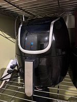Black air fryer with silver handle placed on a wire shelf with visible power cord.