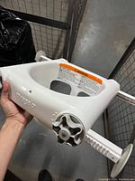 Front angled view of white plastic baby bathtub seat showing adjustable locking arms, circular adjustment grips, and warning label.