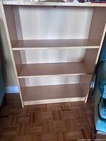 Full view of a light brown wooden bookshelf with three shelves and open back against wall, showing the missing stop on the top shelf and overall condition.