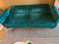 Green velvet futon sofa bed in sofa position on a woven rug, viewed from front and angled slightly right.