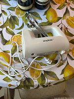 White Simplicité hand mixer with two metal beaters on a yellow lemon patterned tablecloth, with power cord visible.
