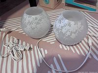 Two frosted glass globe lamps with white floral pattern, placed side by side on striped fabric. Each lamp has attached white power cord with plug and switch visible.