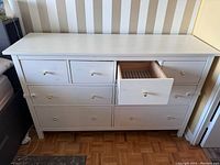 Front view of white IKEA drawer unit with all drawers closed except one drawer open, showing interior and brass-colored knobs.