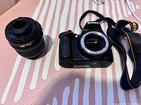 Nikon DSLR camera body with lens mount visible, Nikon branded strap beside it, and detached lens placed adjacent