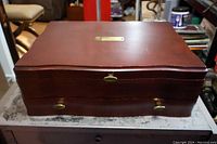 Closed reddish-brown wooden chest with brass handles and a small nameplate on the lid, showing the exterior storage case for the cutlery set.
