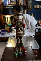 Front view of the 24 inch nutcracker soldier holding drumsticks with gift boxes at the base, set indoors with a white cloth background.