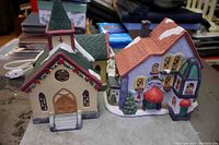Front view of two ceramic Christmas village houses, a church and a toy shop, with light cords visible behind, placed on a table.