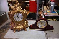 Two clocks, one wood case with brass trim and one ornate gold painted ceramic clock, displayed side by side on a table.