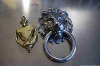 Two door knockers side by side, one a large silver-tone lion head with ring, the other a smaller polished brass traditional style knocker.