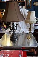 Two table lamps on a metallic surface, one bronze finish globe and arrow design lamp with a brown silk shade, and one solid brass adjustable lamp with a beige fringe silk shade.