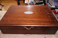 Closed wooden box containing the silverplate cutlery set, showing the brass handle and oval metal label plate on top.