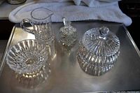 Wide angle photo showing entire 7 piece crystal lot on metal tray, including jug, covered bowl, candy bowl, small square covered piece with spoon.