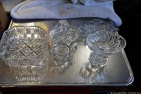 Five crystal items on metallic surface: square bowl, creamer, basket, compote, pressed glass dish