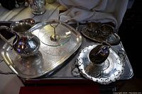 Wide view of silver plated lot showing tray, water jug, glass tidbit tray, cream and sugar set, sugar scuttle