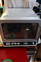 Side/front view of compact wine fridge with glass door and silver border, sitting on a red surface.