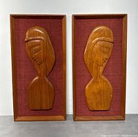 Front view of both teak wood Mid Century Modern abstract female figure wall plaques side by side, showing the relief carving detail and red fabric background within teak frames.