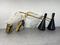 View of the pair of Mercana wall sconces laid flat, shows black conical shades wrapped in plastic and gold-tone arm.