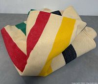 Folded view of the wool blanket showing the colored stripes (green, red, yellow, black) on cream background.