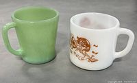 Photo showing both mugs side by side on table, white Beatles mug with brown print and green Fire King Jadeite mug.