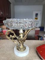 Clear glass faceted bowl ashtray with decorative brass/golden tone metal handle and stem, mounted on white marble base. Mid-century modern design with detailed metal work and solid marble base.