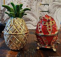 Photo showing two vintage trinket boxes on a wooden surface, one pineapple shaped with green enamel top and rhinestones, the other red royal-inspired egg shaped with red rhinestones and gold-tone base.