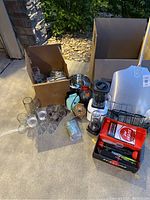 Full assortment of kitchen items and appliances shown outside on pavement including box with glassware, utensil holders, shovel, and various bottles.