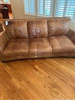 Front view of brown leather sofa showing three seat cushions and back cushions, nailhead trim on armrests, leather in good used condition.