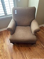 Front view of oversized brown upholstered armchair with rolled arms and wooden legs on hardwood floor.