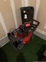 Photo showing the full Toro cordless lawn mower with attached grass collection bag, battery, and charger placed on artificial turf against a beige wall.