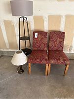Photo showing two patterned upholstered chairs, a tall black metal floor lamp with a gray lampshade and built-in shelf, and a small table lamp with a hexagonal base and beige shade.