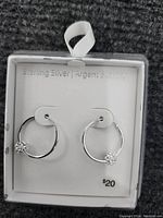 Close-up photo of a pair of sterling silver hoop earrings with a single crystal accent on each. Earrings are in white display packaging marked 'Sterling Silver | Argent Sterling',' priced at $20'.