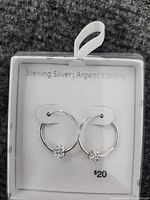 Photo shows a pair of sterling silver hoop earrings displayed side by side in packaging branded 'Sterling Silver | Argent Sterling'. Earrings feature a small floral or cluster detail on one side of each hoop.