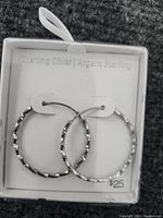 Pair of sterling silver hoop earrings with textured pattern displayed in a white box labeled with Sterling Silver|Argent Sterling and priced $25.