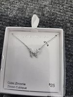 Photo shows a sterling silver butterfly-shaped pendant necklace with cubic zirconia stones on a silver chain inside a white jewelry box labeled 'Sterling Silver / Argentium Silver'. Price tag visible: $25.