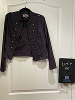 Front view of black gothic military style short jacket with front zipper and decorative button rows, hanging on black hanger against white door.