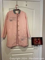 Front view of pink cloth coat with grey collar trim and printed writing, hanging on a red hanger against a white door.