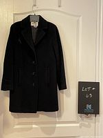 Full front view of a black button-up soft cloth coat hanging on a hanger against a white background, showing its length and overall shape.