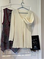 Front view of cream colored knit button-up sweater and multicolored crocheted sleeveless pullover coverup hanging on hangers