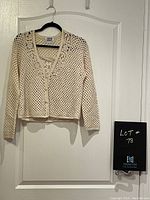 Full view of beige crocheted cardigan hanging on a black hanger against a white door.