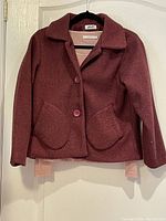 Dark coral short lined blazer on hanger showing front with collar, buttons, and two rounded patch pockets. Pink pullover partially visible underneath.