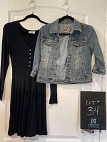 Light-wash denim jacket and black V-neck dress hung side by side on hangers against a white door, clearly showing front details of both garments.