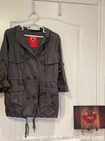 Front view of the lightweight grey EDC car coat hanging on a white hanger against a white door, showing the hood, double-breasted button design, and pockets.