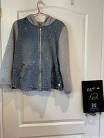 Front view of the hooded zip sweatshirt hanging on a hanger on a white door. Shows denim front panel with distress marks, grey sleeves and hood with print details, zipper, and side pockets.