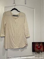 Front view of large light beige women's top with faux pearl decorations, hanging on a black hanger on a white door.