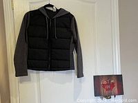 Front view of Whisper brand jacket hanging on black hanger showing black padded front panel and grey jersey sleeves and hood