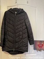 Front view of the black long quilted jacket with hood and front zipper, hanging on a hook.