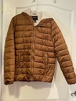 Front view of men's dark tan quilted bomber jacket hanging on white hanger, showing full zipper, front pockets, and hood.