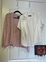 Pink zip-up cardigan and white short sleeve cotton shirt hanging on hangers on a door.