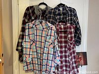 Four plaid shirts hanging on a door in various colors and styles including long-sleeve and sleeveless.