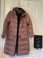 Front view of light purple maxi length puffy winter coat with large pockets, double zipper, and hood hung on door.